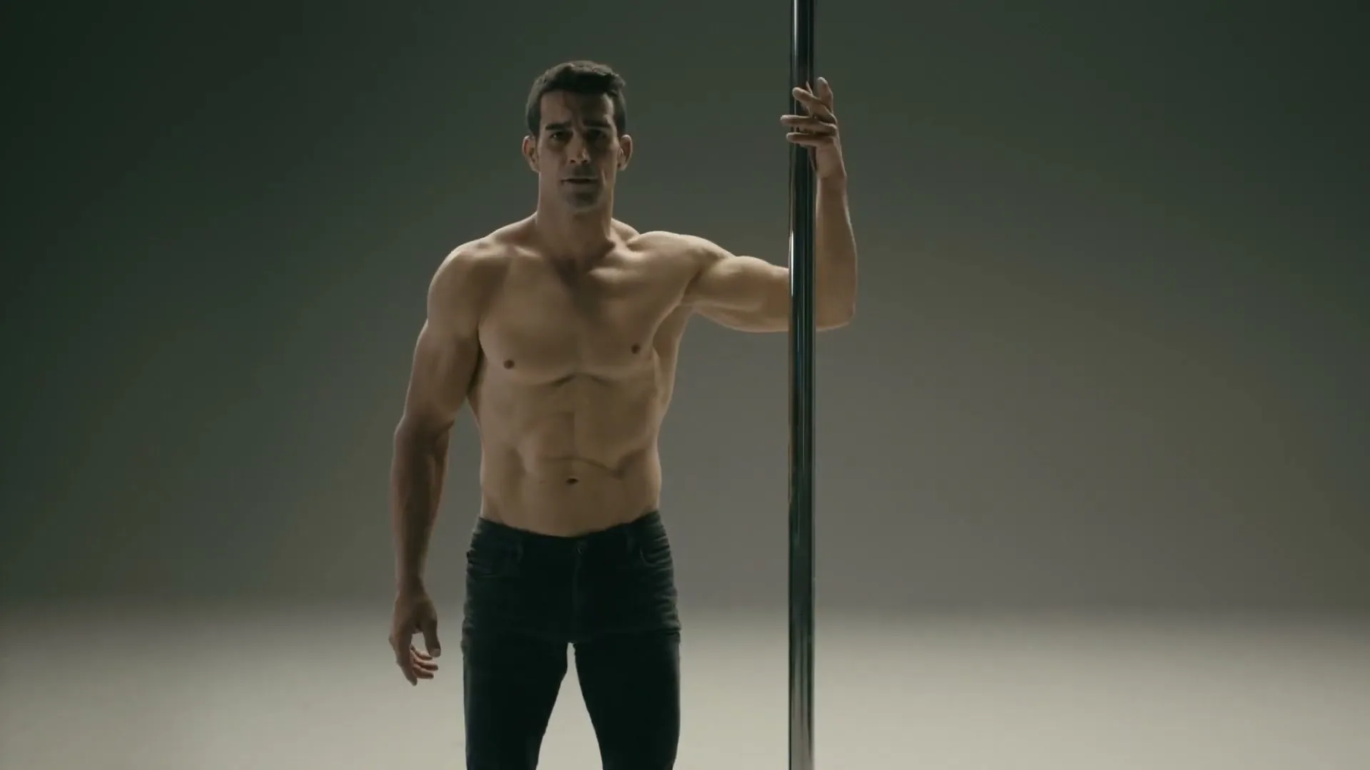 A muscular man in his 30s, shirtless and wearing black pants, stands next to a pole in a studio, looking at the camera with a neutral expression.