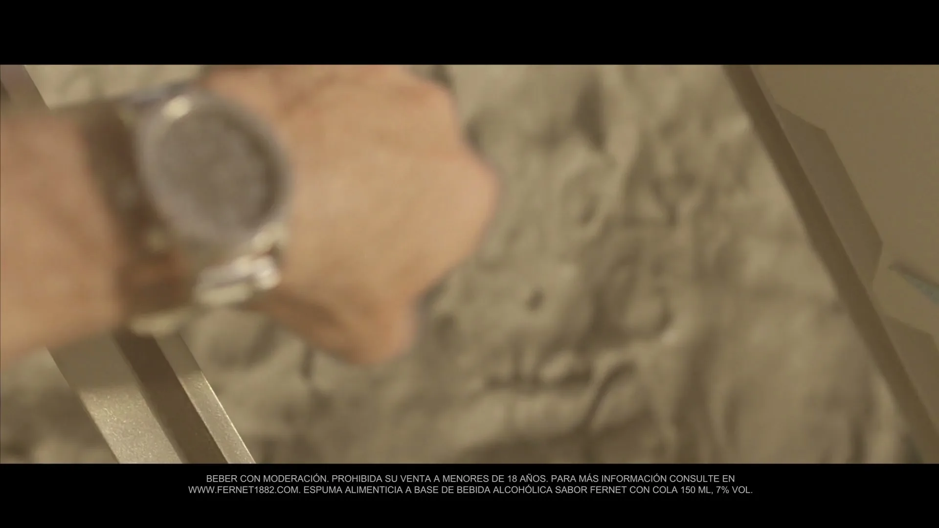 A man's arm with a watch is seen in a medium closeup, as he reaches for something in a dimly lit, indeterminate interior setting.