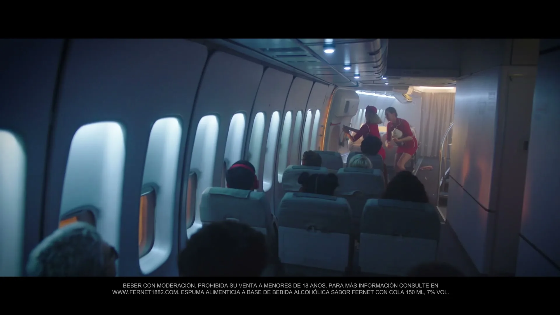 Two women in red dresses walk down the aisle of an airplane, carrying drinks, as passengers sit in their seats.
