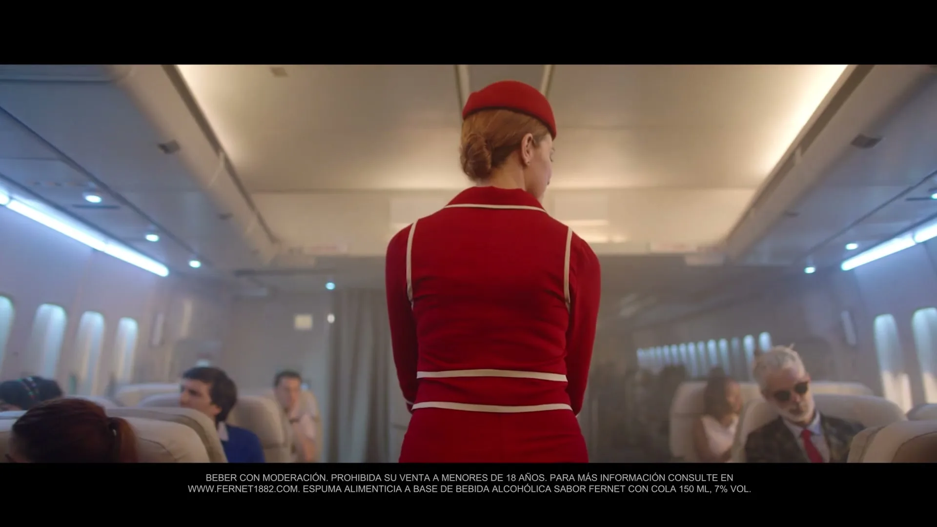 A female flight attendant in a red uniform stands in the aisle of an airplane, looking back at the passengers.