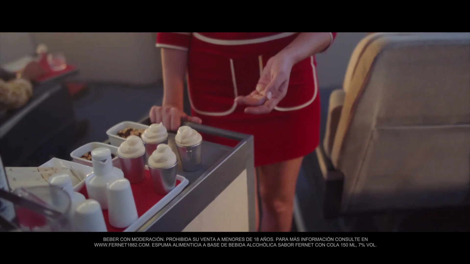 A female flight attendant in a red uniform pushes a beverage cart with small cups of whipped foam on an airplane.