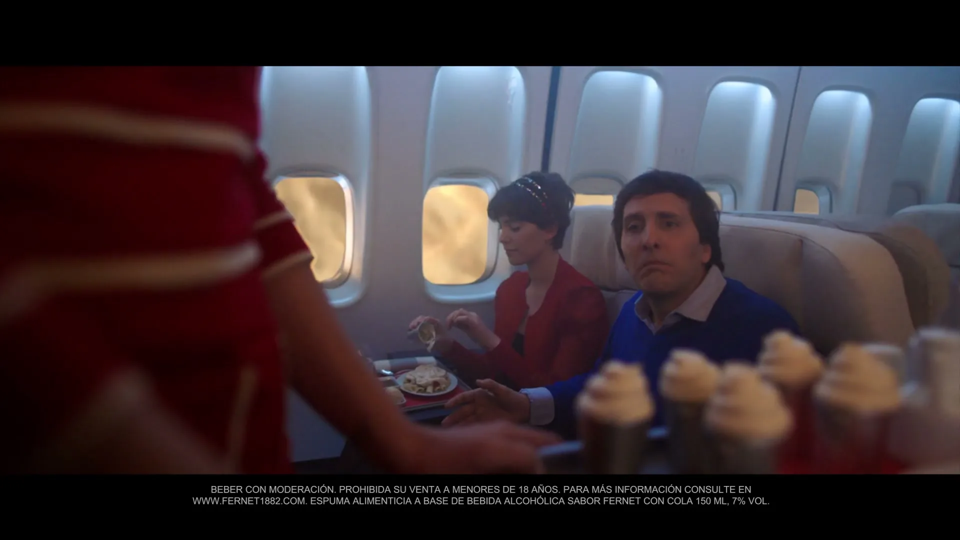A flight attendant in a red uniform serves drinks to a male and female passenger on an airplane, with bright sunlight streaming through the windows.