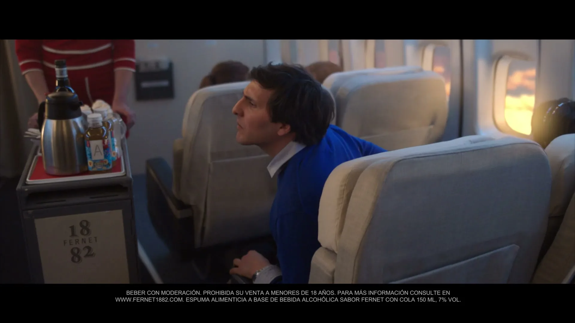 A man in a blue sweater looks up from his airplane seat as a flight attendant pushes a cart with drinks down the aisle.