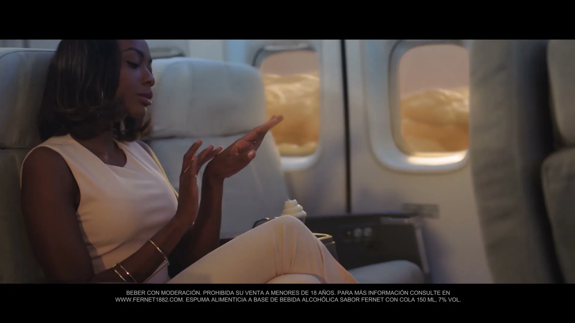 A woman in her 20s, wearing a light pink dress, sits in an airplane seat, looking at her hands as if holding something, with clouds visible outside the window.