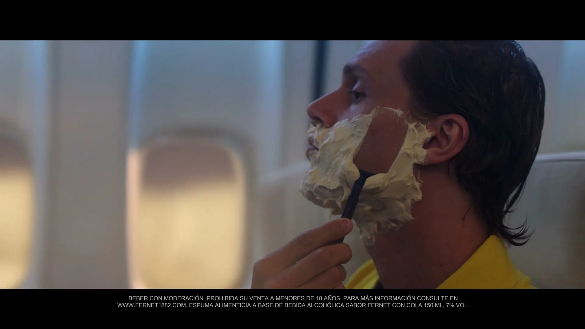 A man with shaving cream on his face shaves with a razor on an airplane, looking out the window.