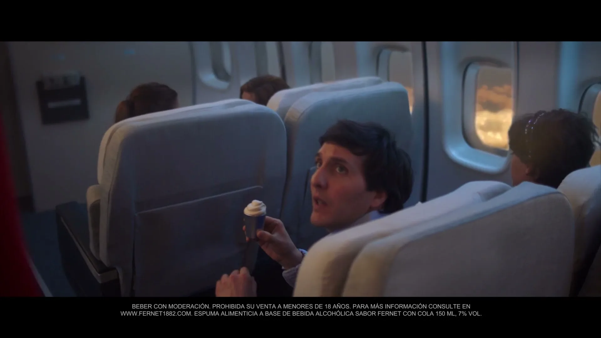 A man in his 30s, wearing a blue shirt, holds a small ice cream cup and looks up at a flight attendant in a plane cabin.