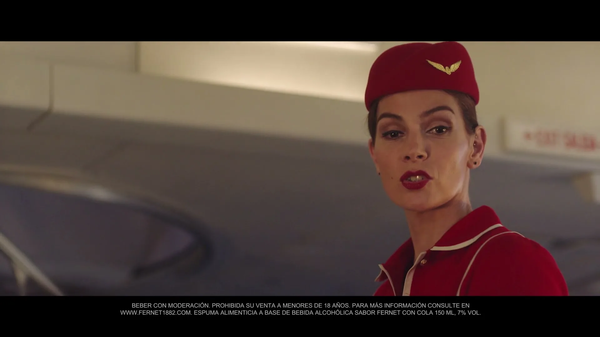 A female flight attendant in a red uniform and hat looks at the camera, then turns her head to the right, inside an airplane cabin.