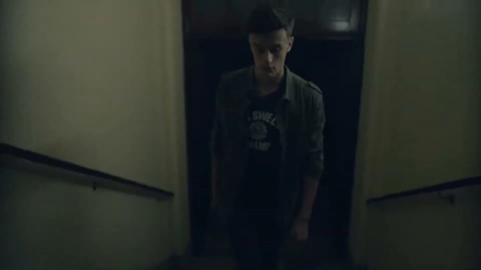 A young man in a dark t-shirt and green jacket walks down a dimly lit concrete stairwell, looking around nervously.