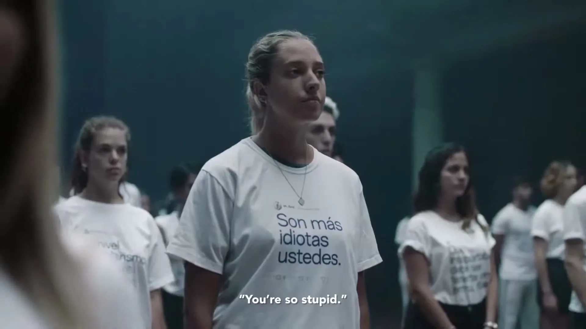 A young woman with blonde hair in a ponytail stands in a large group of people, all wearing white t-shirts with text, in a dimly lit room.