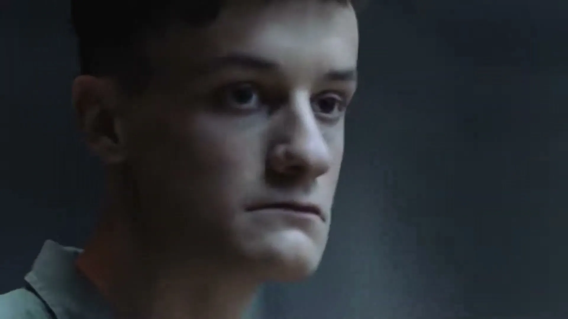 A young man with short hair and a green shirt looks around nervously in a dimly lit, dark room.