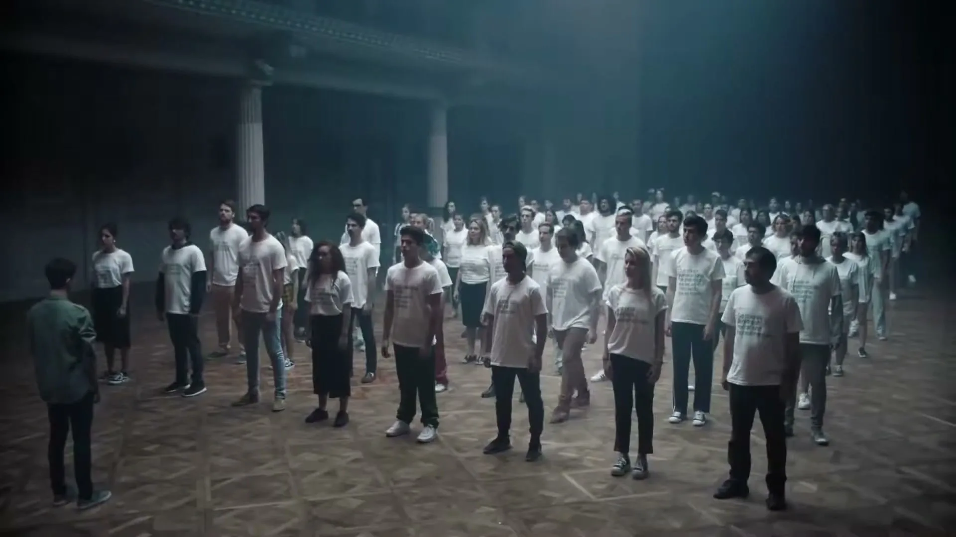 A young man in a green shirt stands facing a large crowd of people in white t-shirts in a dimly lit, empty hall.