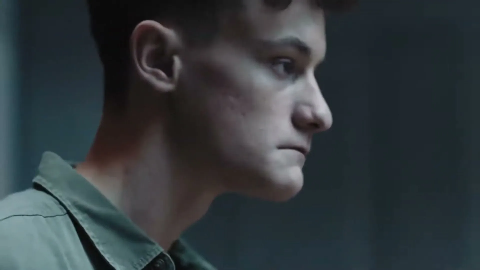 A young man with short dark hair and a green shirt looks down and to the right, with text overlayed on the screen.