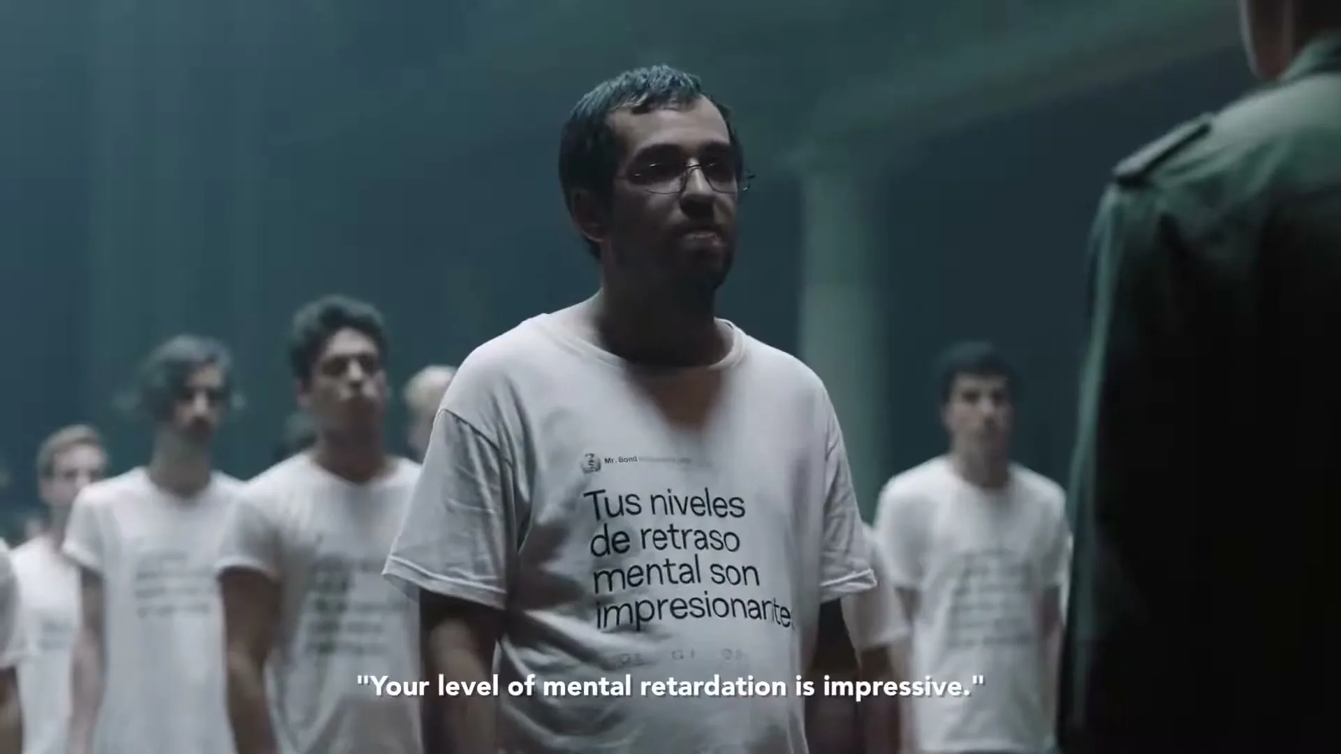 A man in glasses and a white t-shirt with Spanish text stands in a line of people, facing a person in a military uniform in a dimly lit, industrial-looking room.