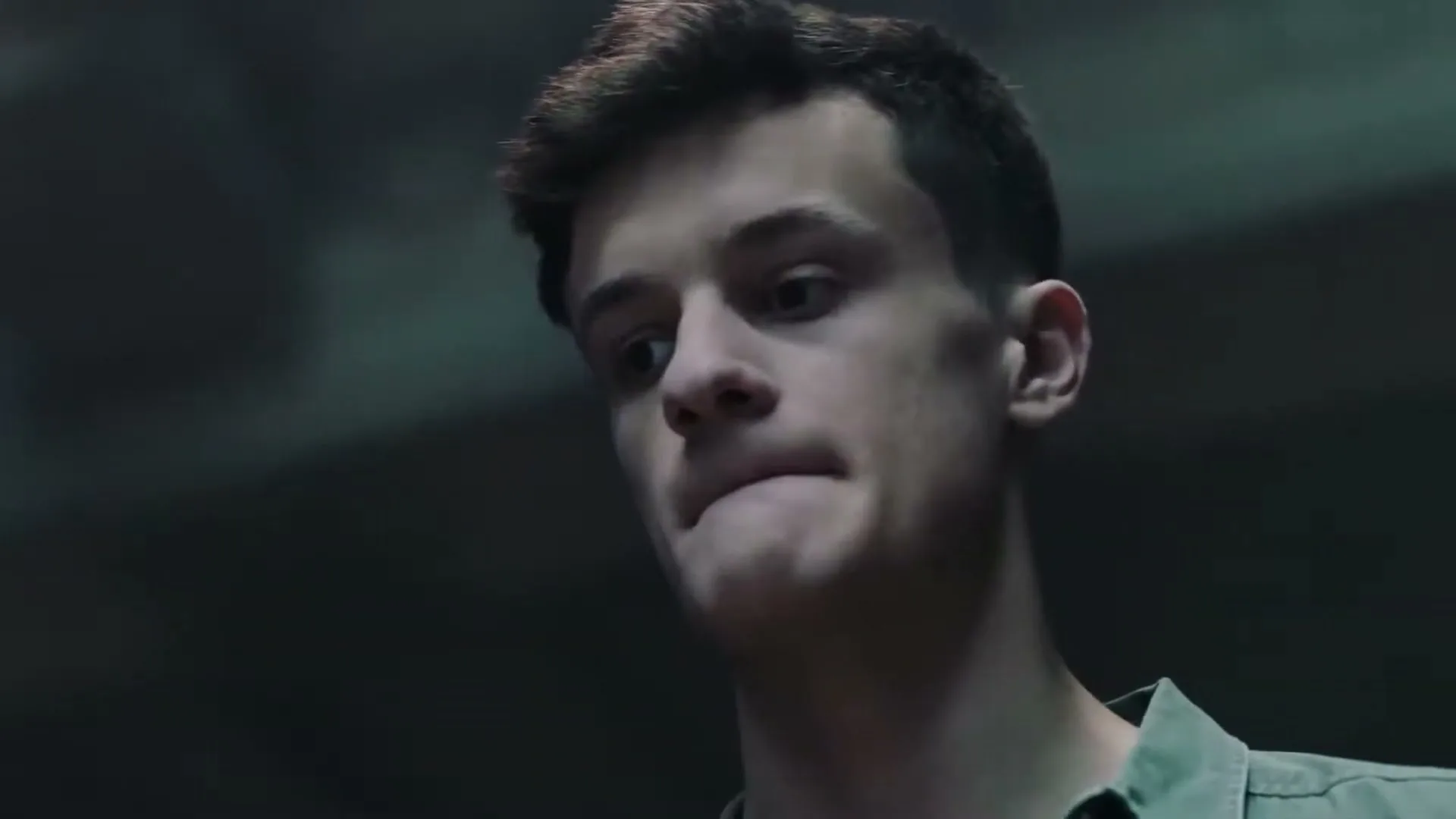 A young man with short dark hair and a green shirt looks down with a pained expression, as text appears on screen in a dark, ambiguous setting.