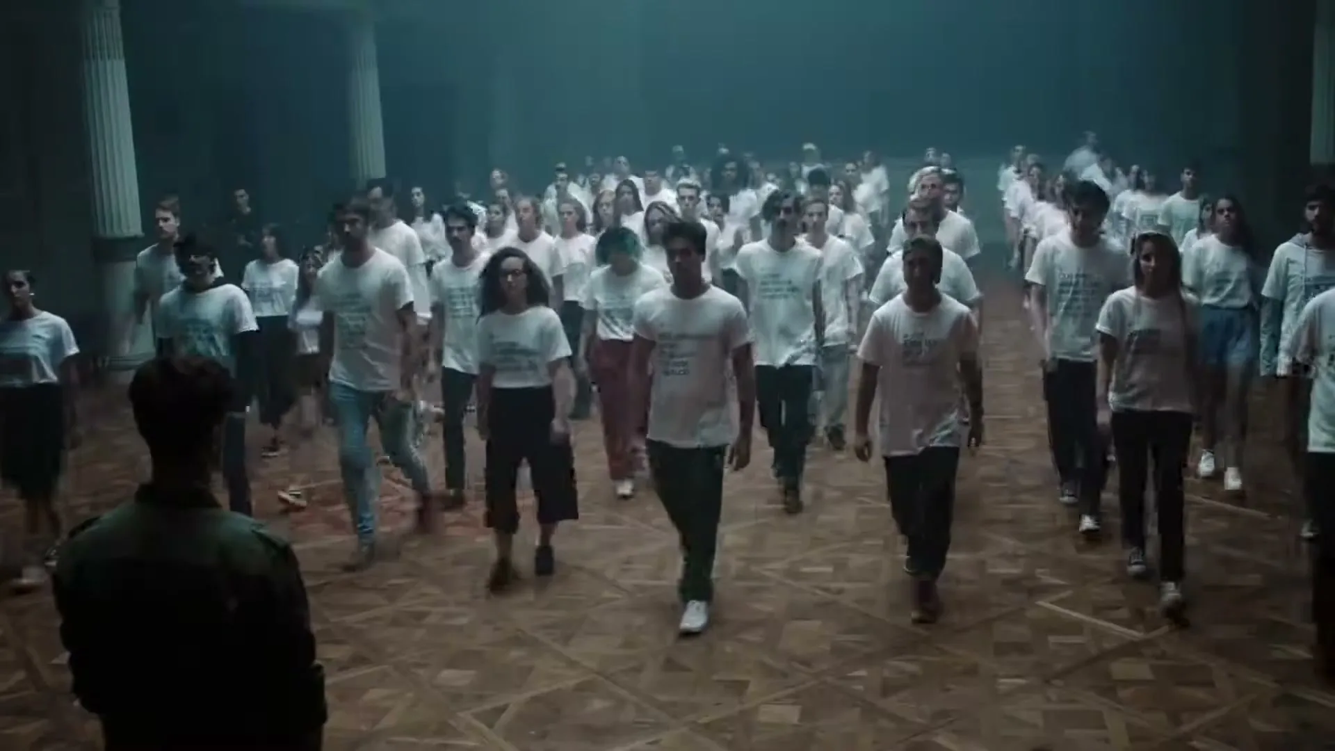 A large group of diverse young adults in white t-shirts walk towards the camera in a dimly lit, smoky hall with columns.