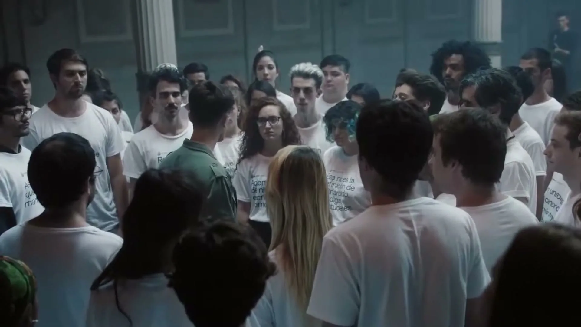 A young man in a green jacket stands in the middle of a large group of young people, all wearing white t-shirts, in a dimly lit hall.