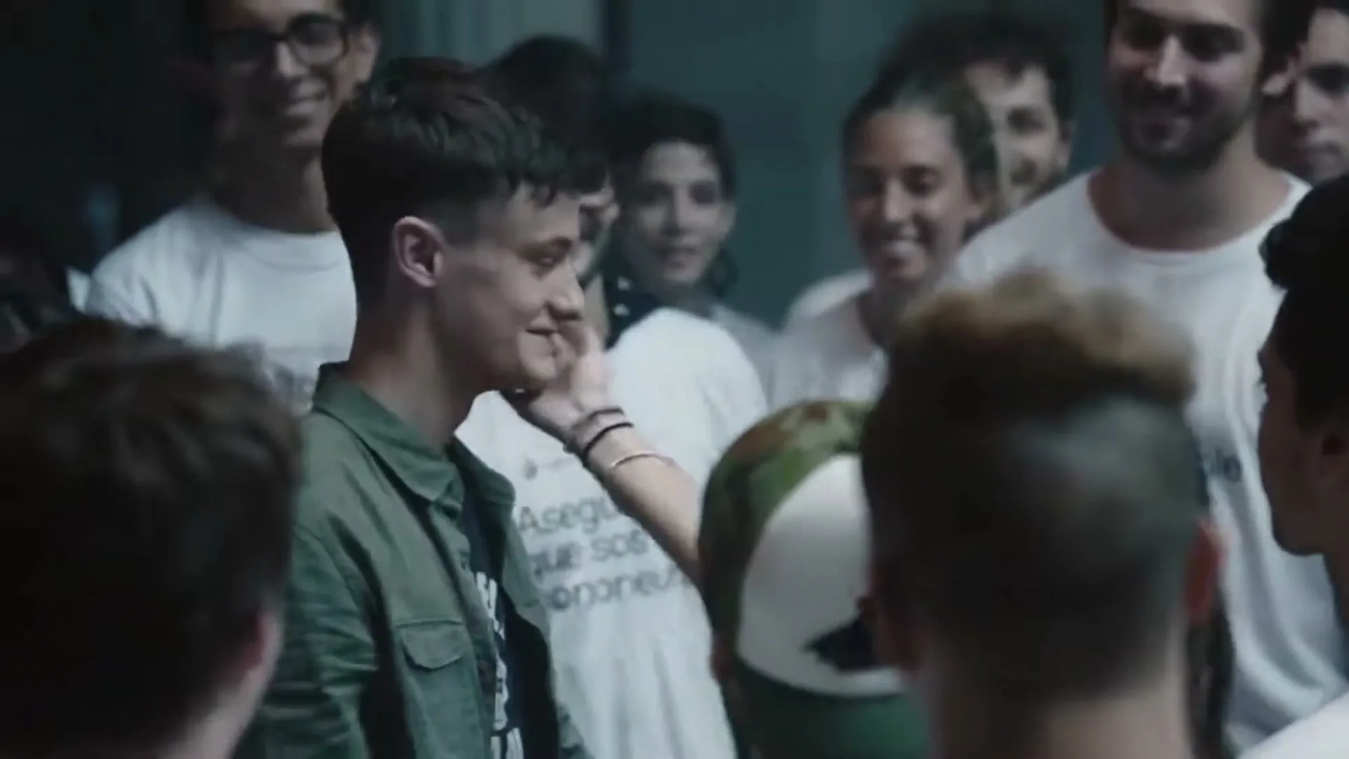 A young man in a green jacket is embraced by a woman in a green shirt, surrounded by a crowd of smiling people in white t-shirts.