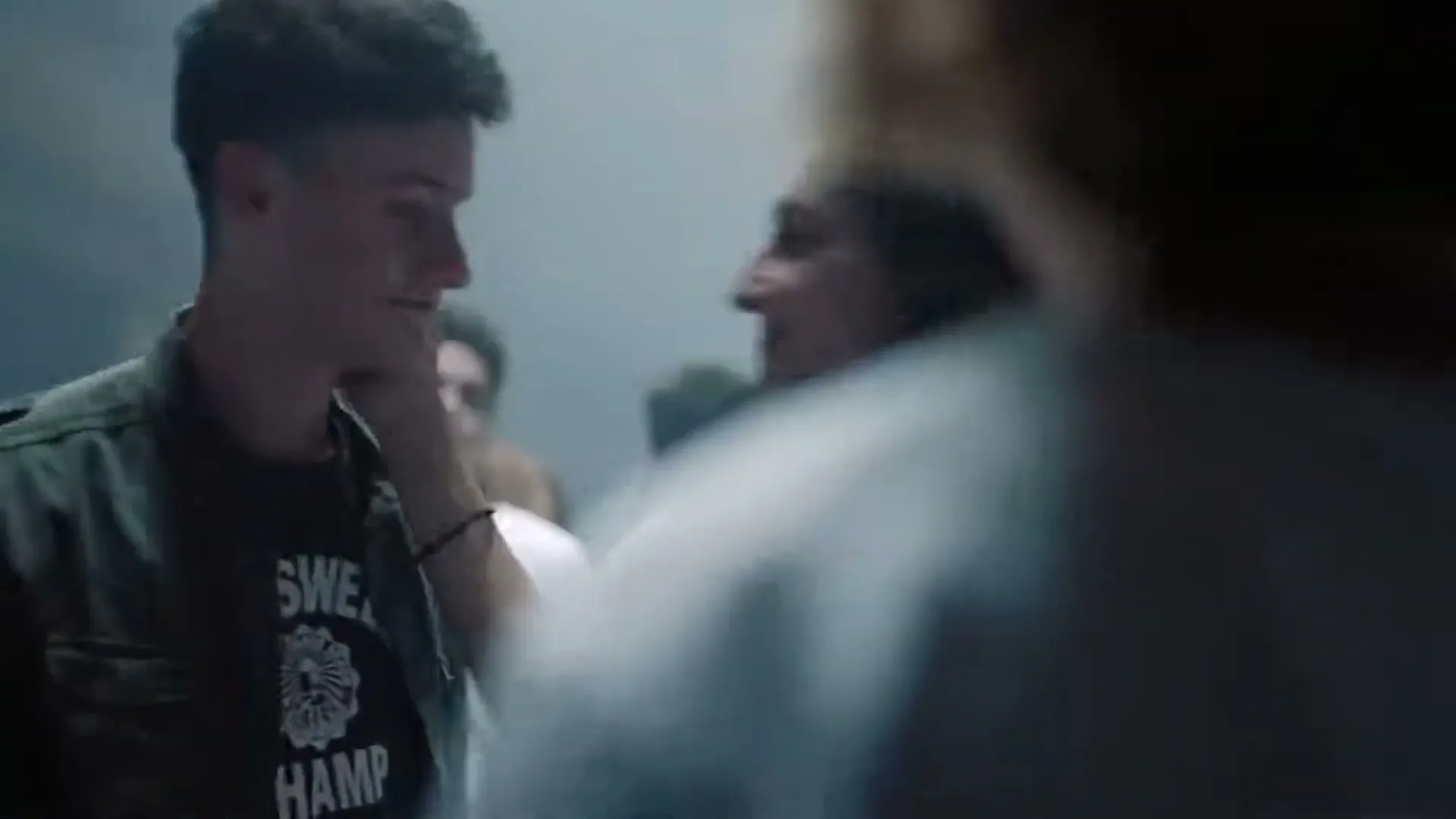 A young man in a green jacket and a young woman with dark curly hair hug each other in a crowd of people, all wearing white t-shirts, in a smoky, dimly lit room.