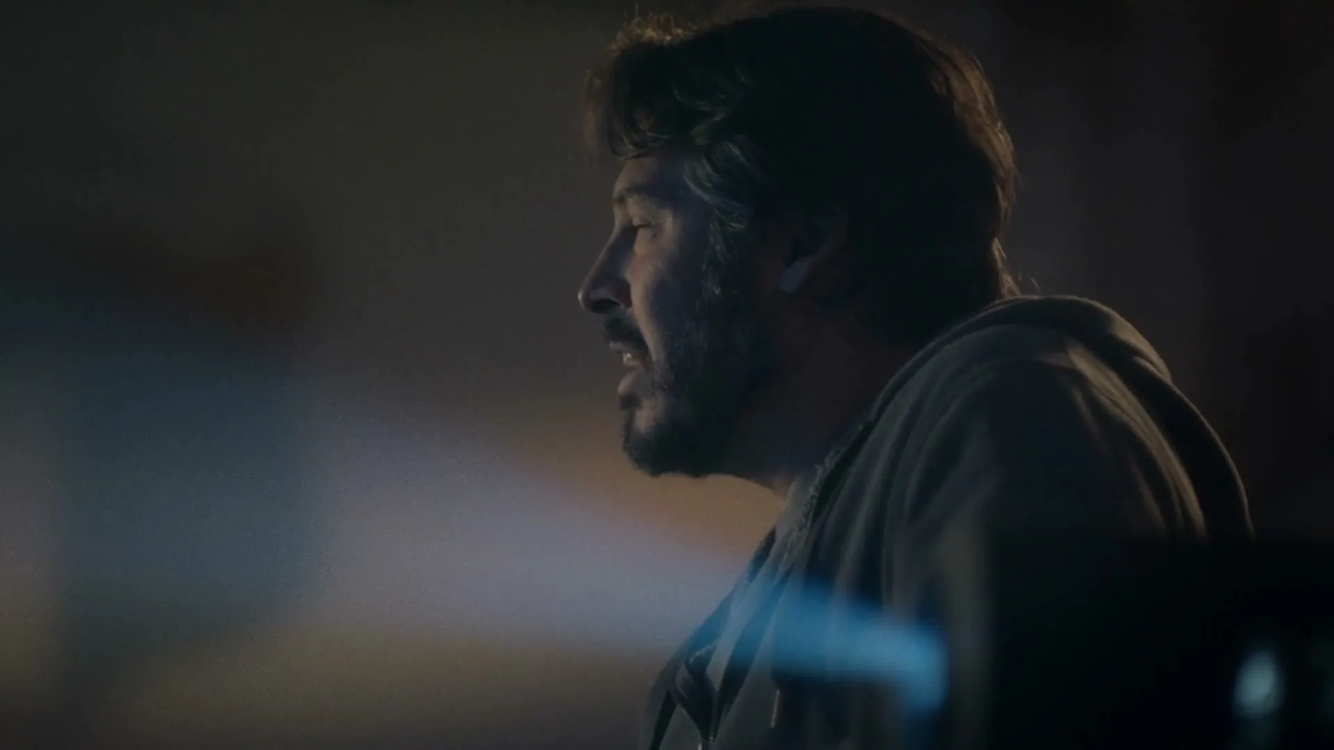 A man with a beard and dark hair, wearing a grey hoodie, looks off-screen with a thoughtful expression in a dimly lit room.