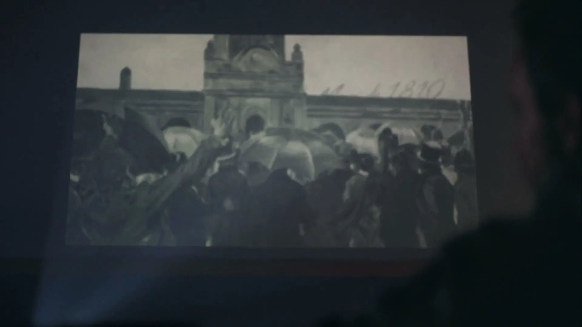 A person's back of the head is visible as they watch a black and white archival projection of a crowd gathered in front of a building.