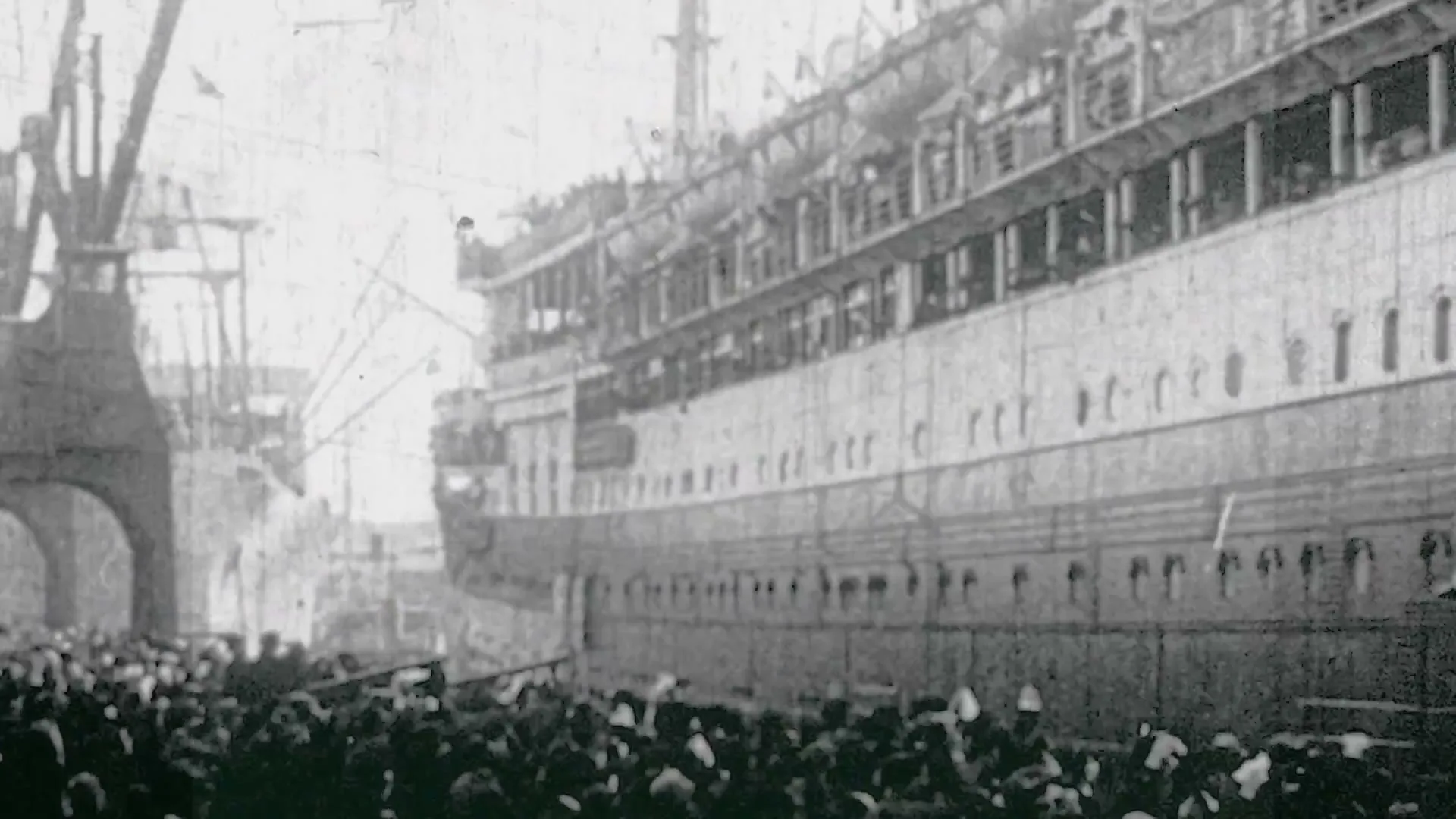 A large crowd of people gathers at a dock to watch a massive ship, possibly a passenger liner, being launched or departing in this archival footage.