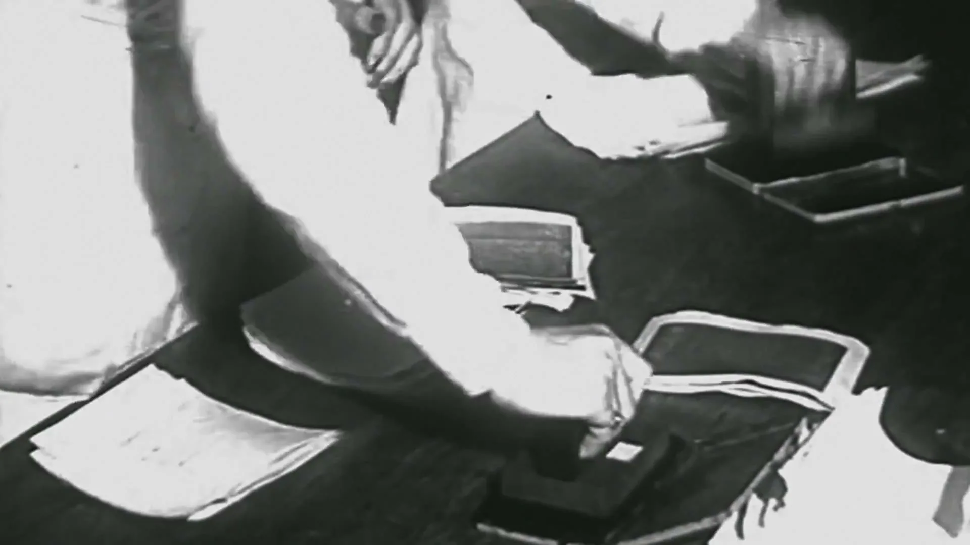 A person in a white shirt stamps documents on a dark table, with stacks of paper and ink pads visible in a black and white, archival footage style.
