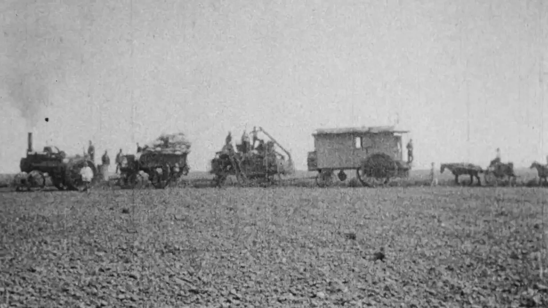 A black and white archival footage shot shows a line of horse-drawn wagons and early agricultural machinery moving across a vast, barren field under a bright sky.