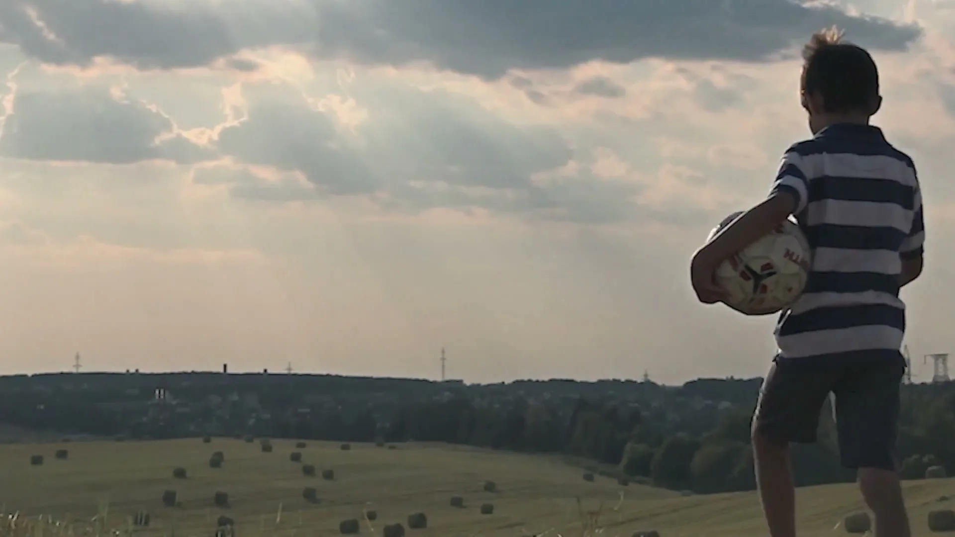 A young boy in a striped shirt holds a soccer ball and looks out over a vast field with hay bales under a cloudy sky.
