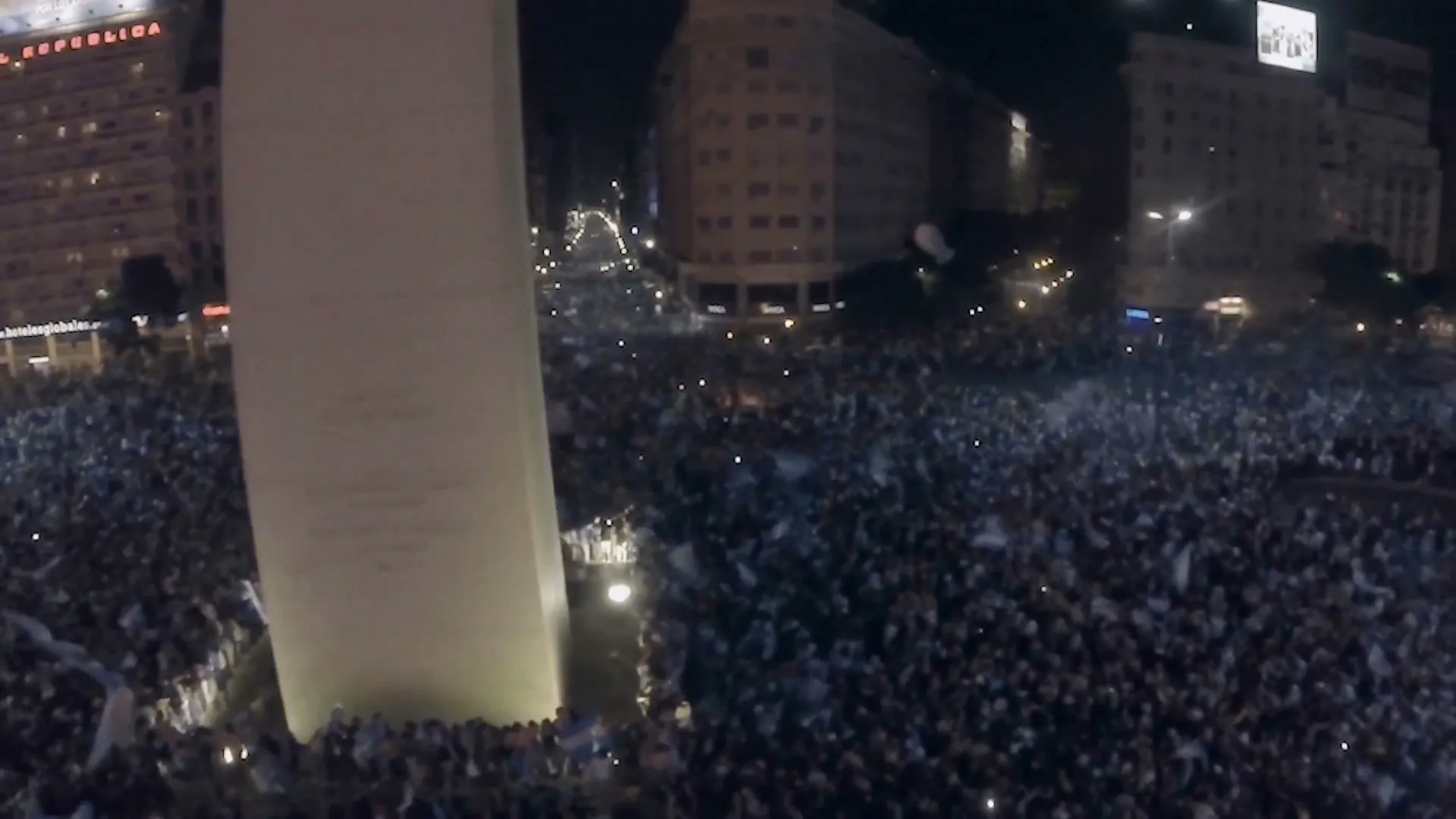 An aerial view shows a massive crowd of people gathered around the Obelisco de Buenos Aires at night, celebrating with flags.