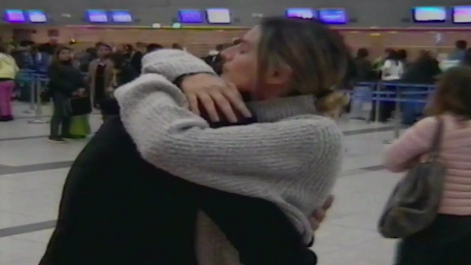 A woman with blonde hair in a grey sweater hugs a man in a black jacket at an airport, with other travelers in the background.