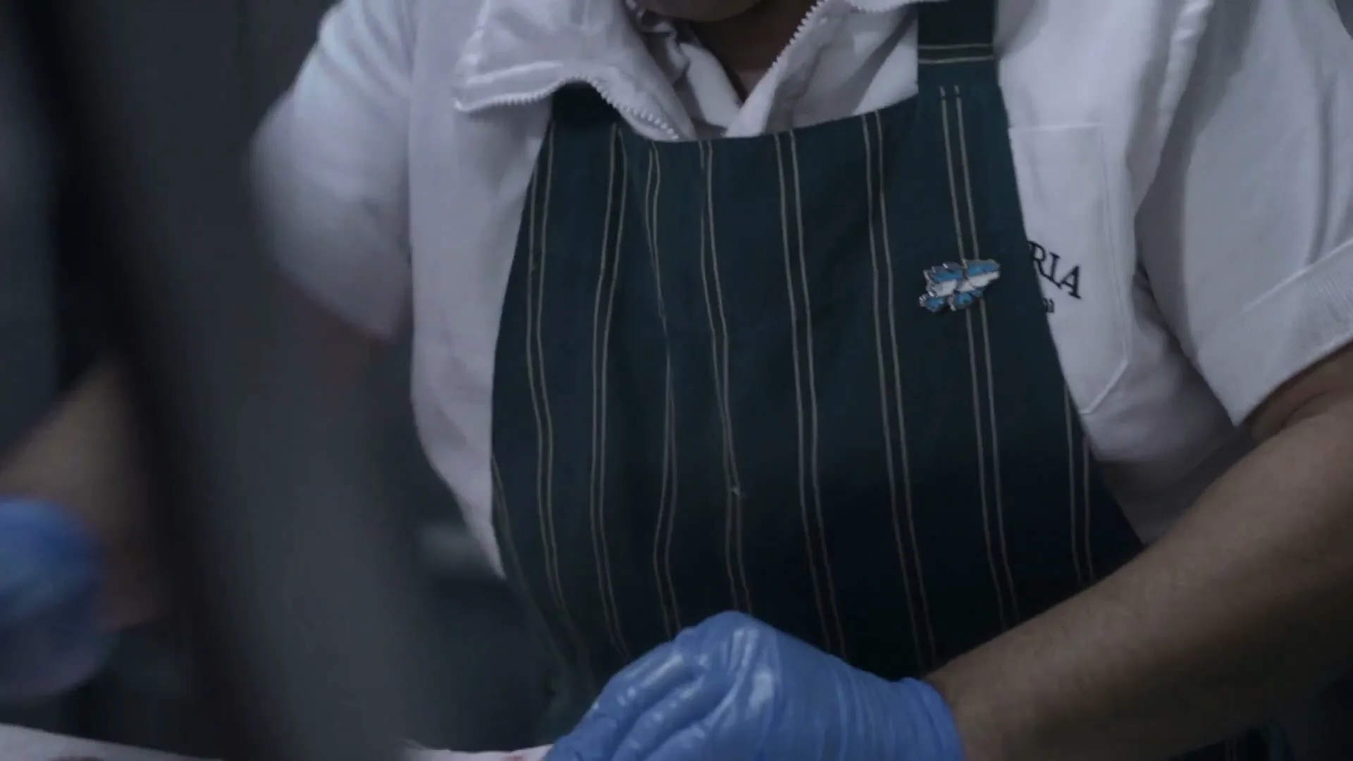 A butcher in a white shirt and striped apron, wearing blue gloves, is seen cutting meat in a dimly lit interior.