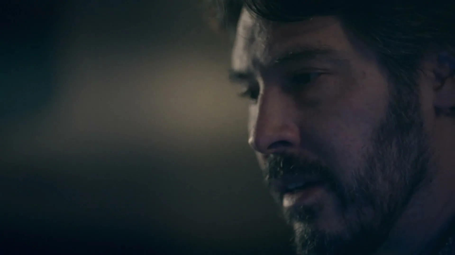 A man with a beard looks down and to the right, his face partially illuminated in a dark, moody setting.