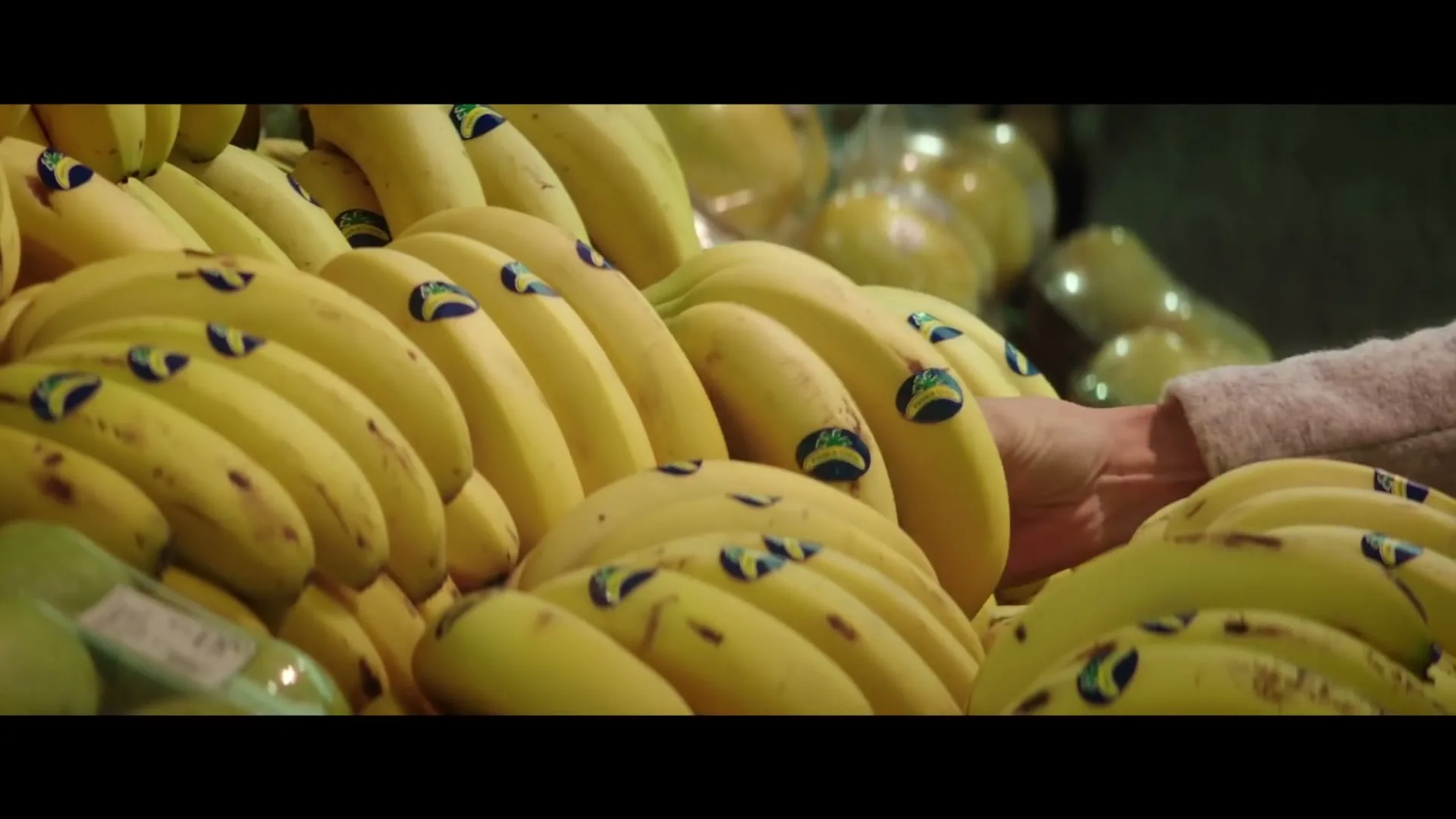 A person's hand reaches into a pile of yellow bananas with blue stickers in a grocery store produce section.