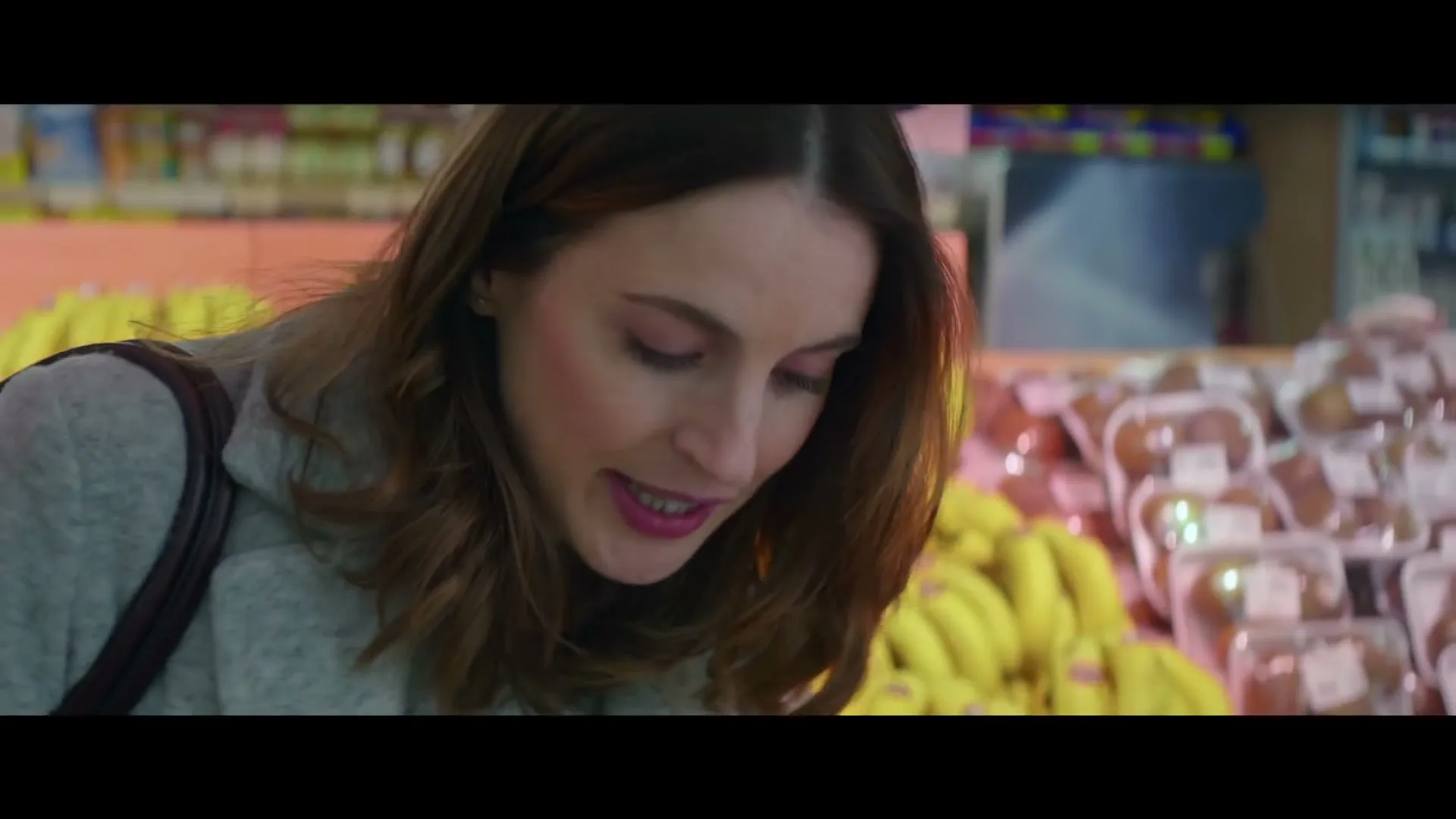 A woman with brown hair and a grey coat looks down at bananas in a grocery store, then looks up and smiles.