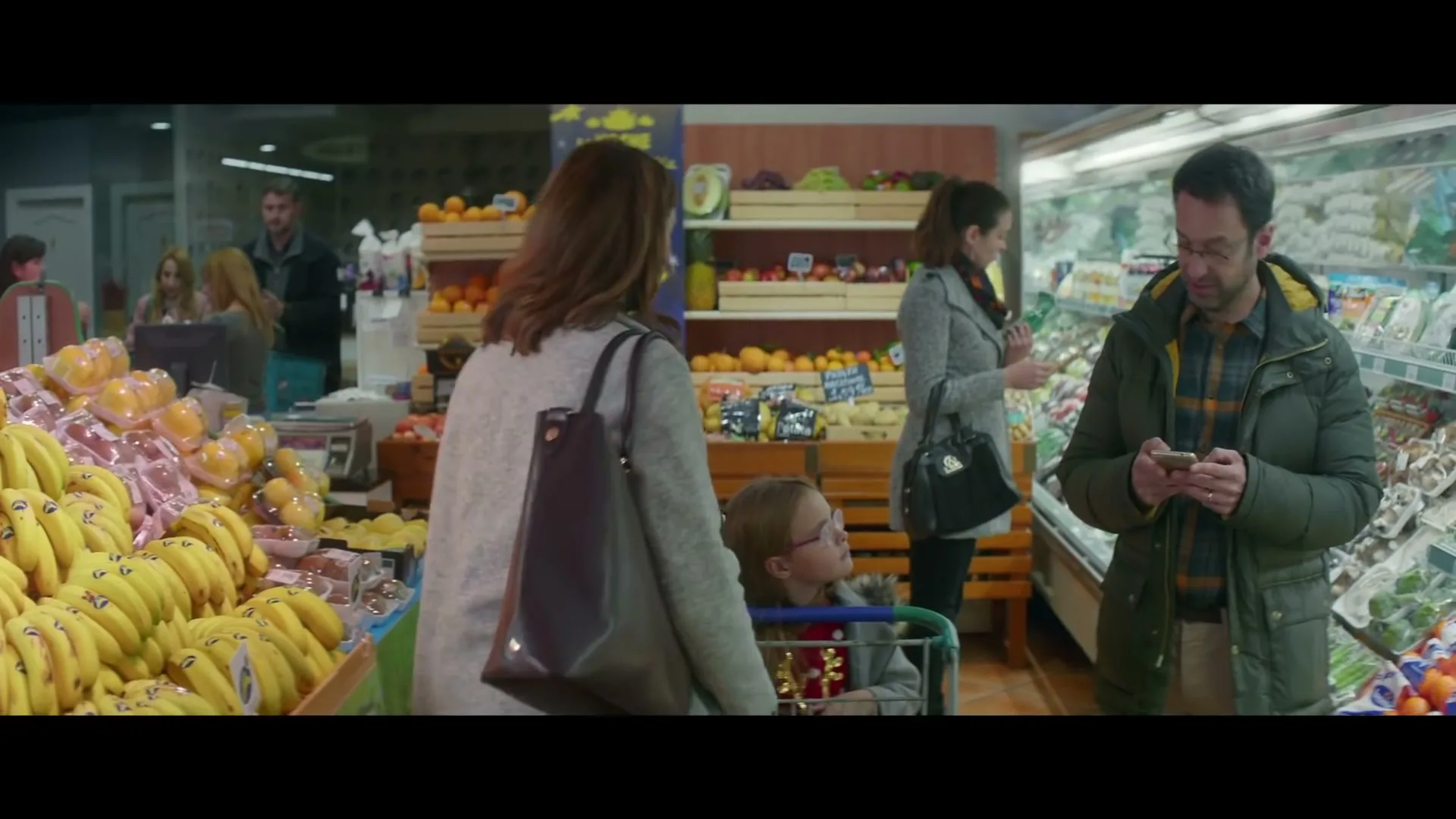 A woman with a child in a shopping cart walks past a man looking at his phone in a brightly lit supermarket produce section.