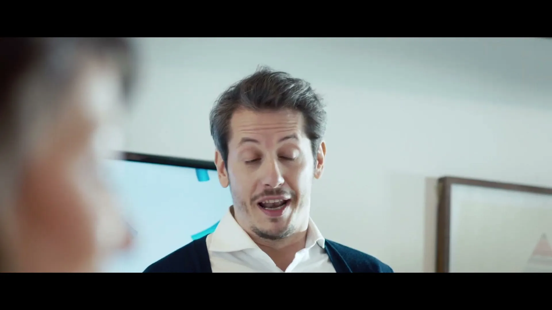 A man in his 30s, wearing a white shirt and dark cardigan, speaks to someone off-camera in a brightly lit room with a TV screen in the background.
