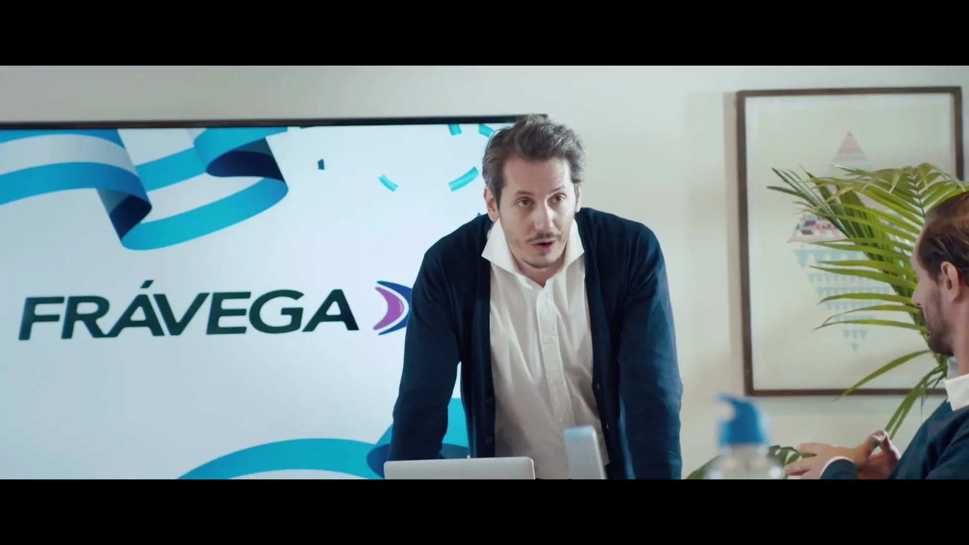 A man in a white shirt and blue cardigan leans on a table, talking to colleagues in a modern office with a large screen displaying a logo.