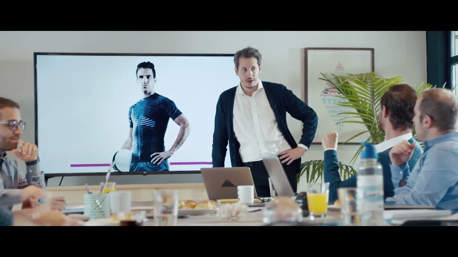 A man in a white shirt and dark cardigan stands in front of a large screen showing a male athlete, addressing a group of people seated around a conference table in a modern office.