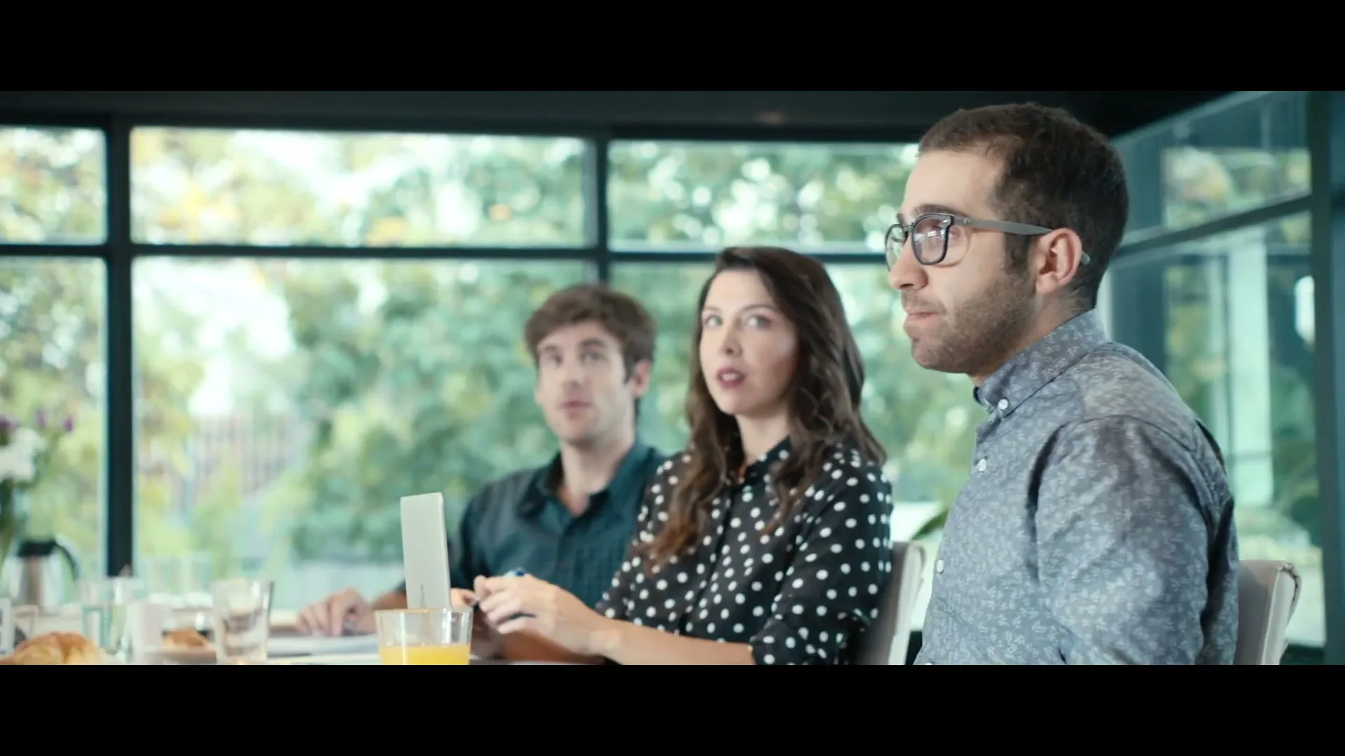 Three young adults, two men and one woman, sit at a table in a modern office with large windows, looking off-camera with varied expressions.