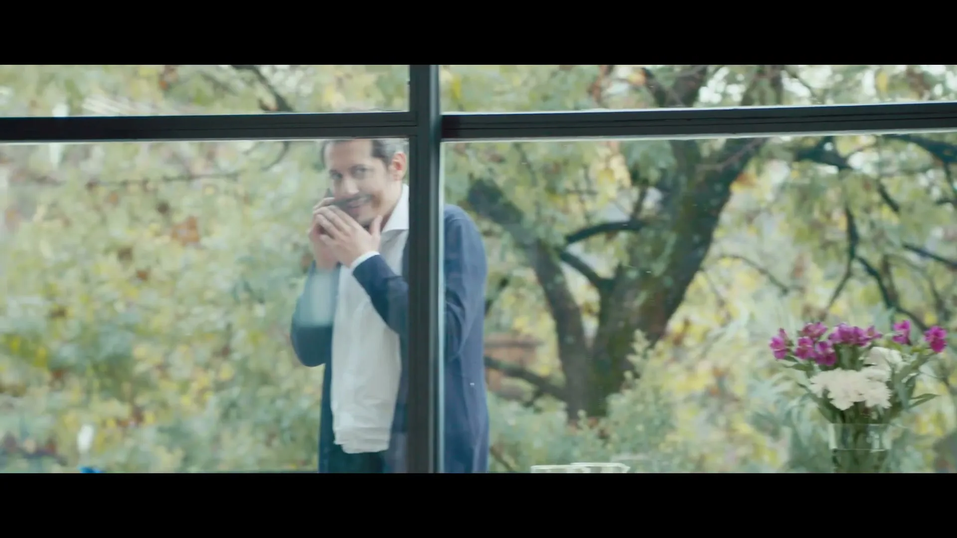 A man in a blue cardigan and white shirt talks on the phone, smiling, while looking out a large window with trees and flowers visible outside.
