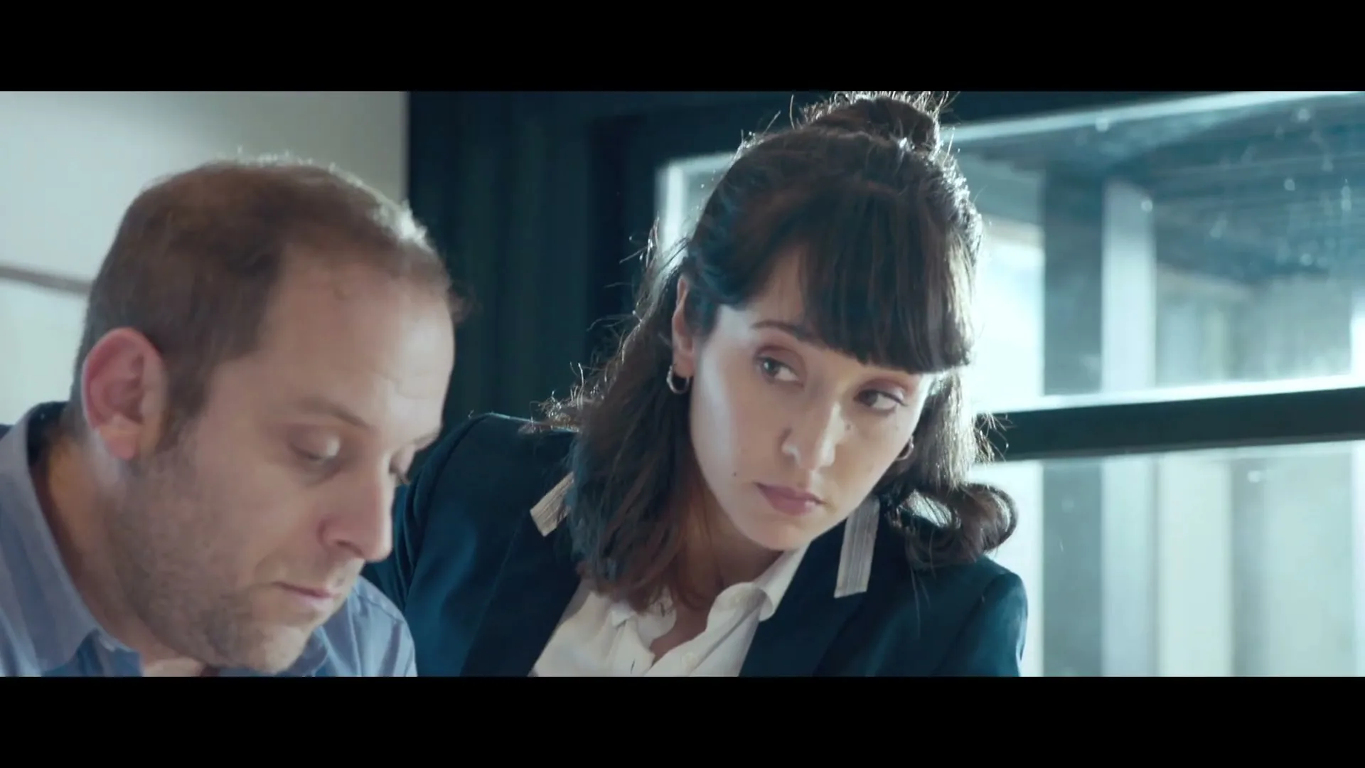 A man and a woman, both in their 30s, look down at something off-screen in an office setting.