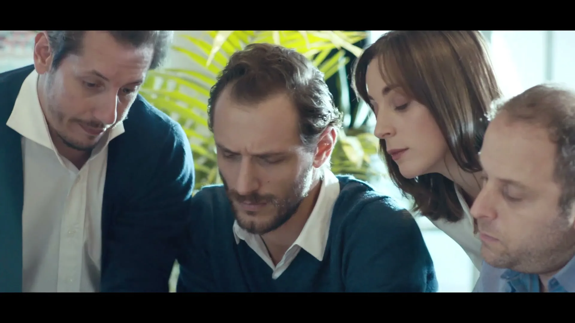 Four colleagues, three men and one woman, lean in closely to look at something on a table in a brightly lit office.