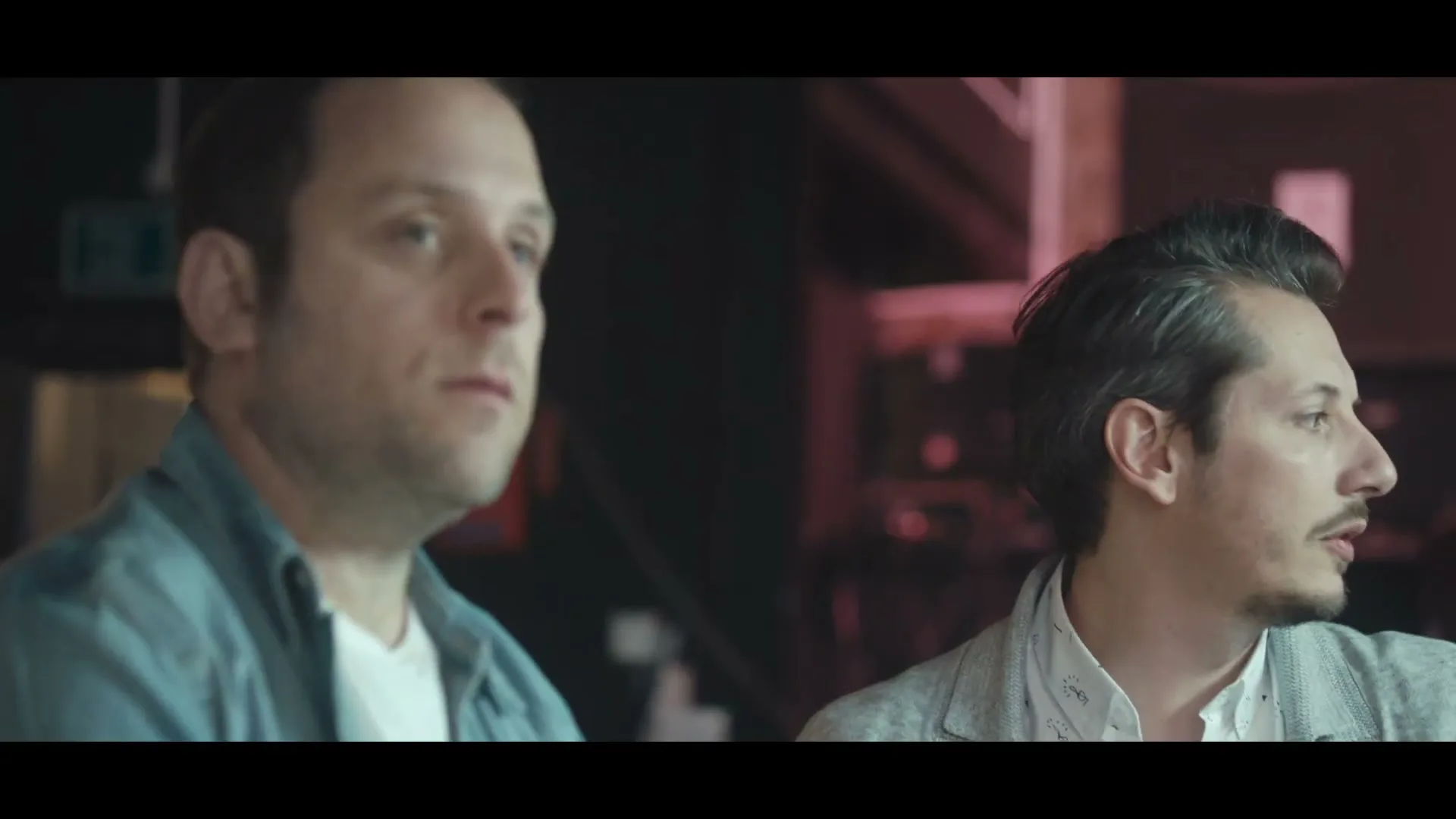 Two men, one in a blue shirt and one in a patterned shirt, look to the right in a dimly lit interior.