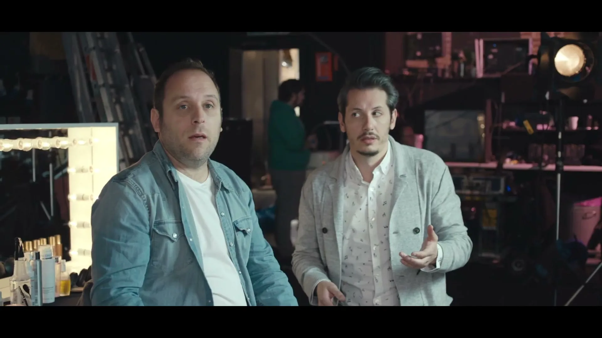 Two men, one in a denim shirt and the other in a patterned shirt and light jacket, look forward with surprised expressions in a backstage area.