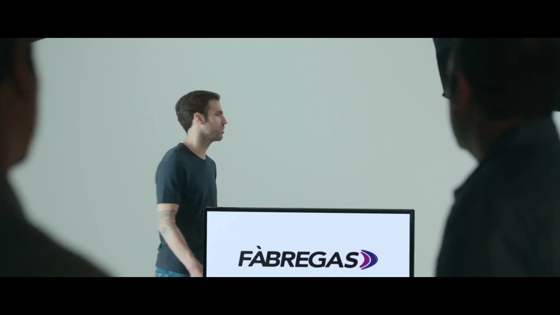 A man in a dark t-shirt stands in a studio, looking at two other men whose backs are to the camera, with a screen displaying a logo in the foreground.