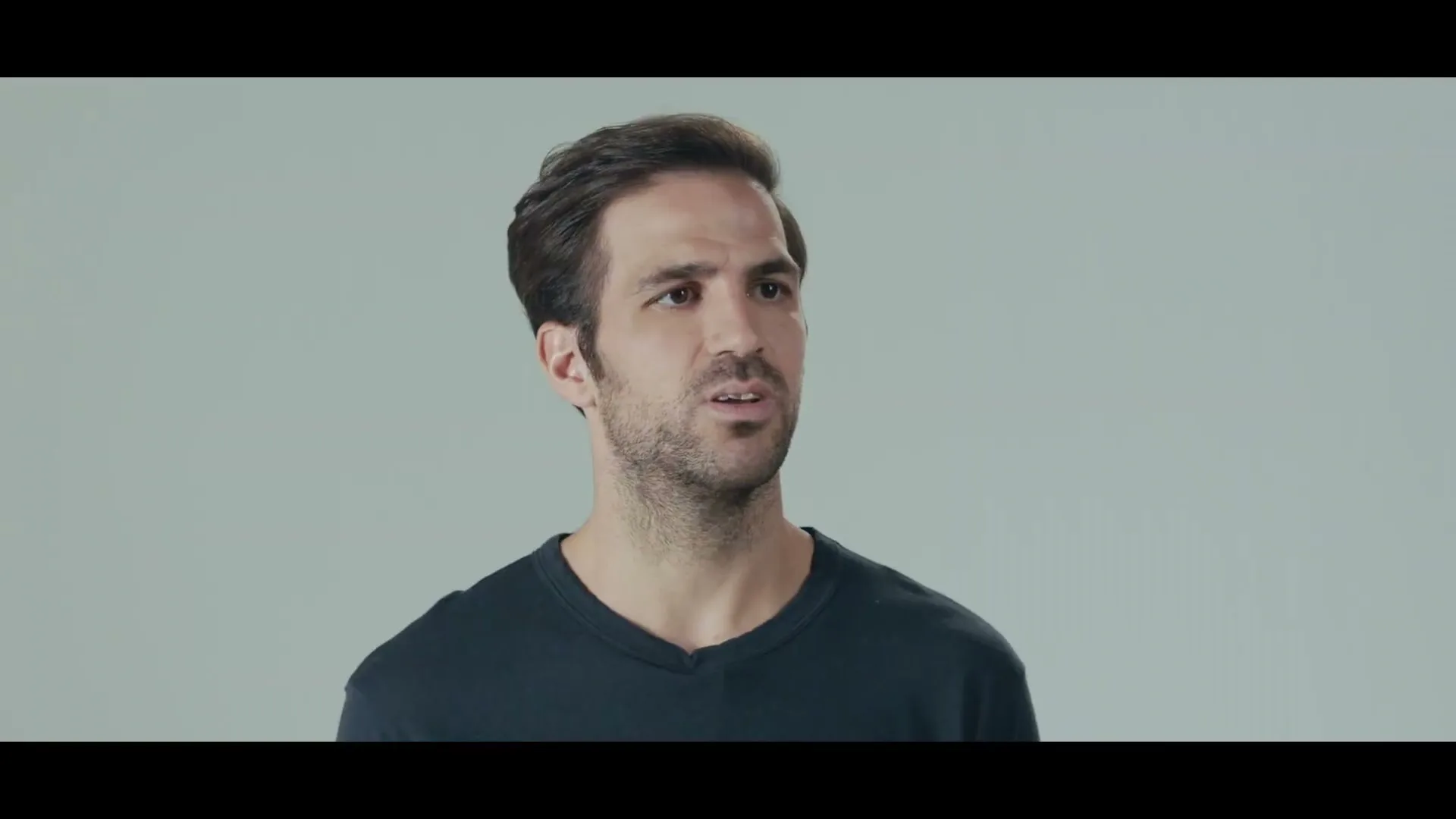 A man with dark hair and a beard, wearing a dark t-shirt, looks up and to the right with a confused expression in a studio.