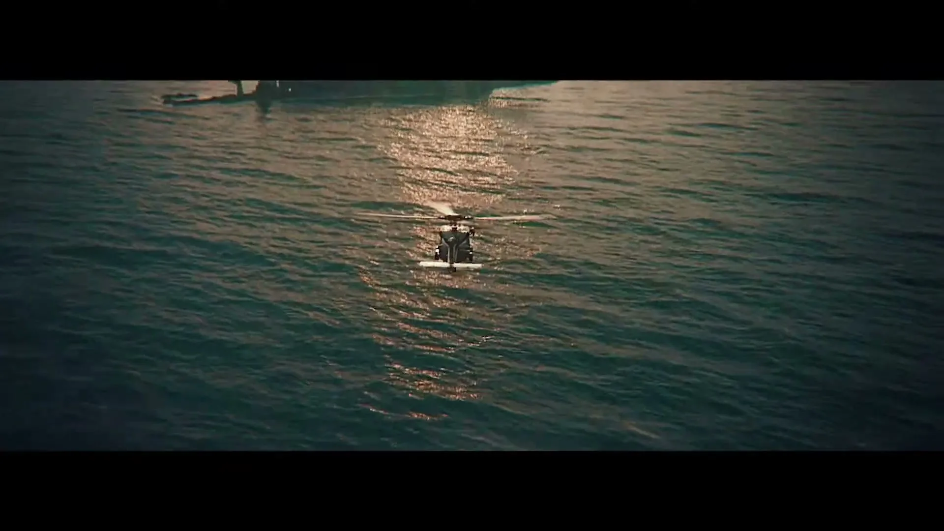 An aerial shot shows a black helicopter flying over the ocean towards a large, dark submarine at sunset.