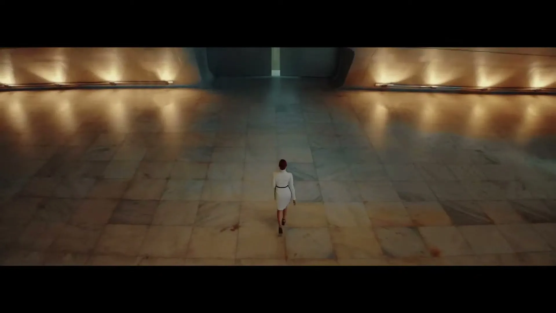 A woman in a white dress walks away from the camera down a long, empty, marble-floored hallway with warm lights on the walls.