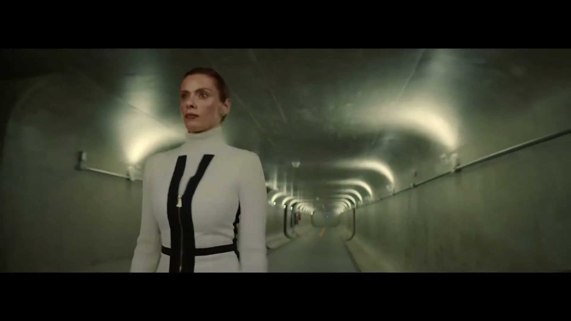 A woman with a stern expression walks towards the camera in a long, dimly lit, futuristic tunnel.