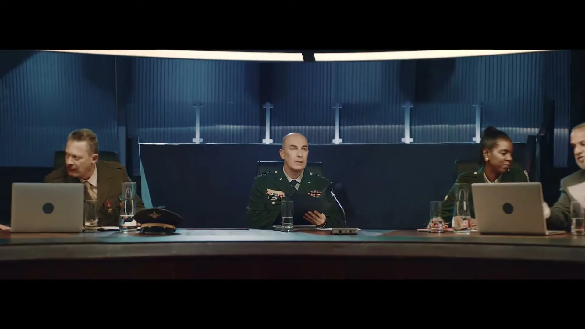 A diverse group of military generals and officials sit around a large, curved conference table in a dimly lit, futuristic meeting room, looking concerned.